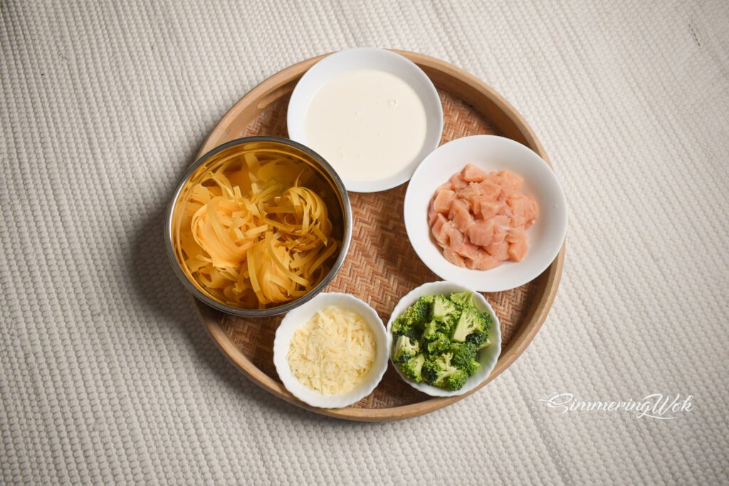 Creamy garlic chicken pasta ingredients arranged on a round wooden tray including fettuccine chicken cream parmesan and broccoli